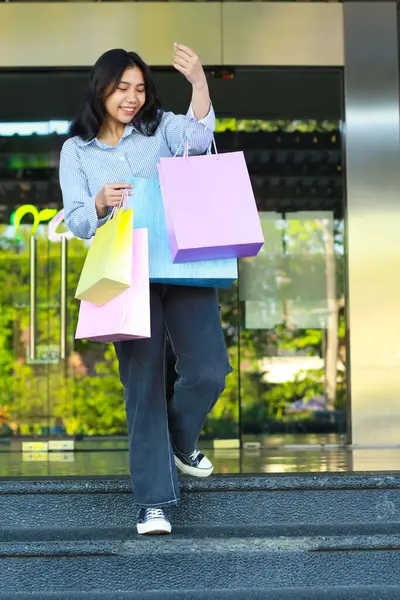 vertical shoot of laughing asian woman holding shopping bag walking on staircase in front of mall entrance