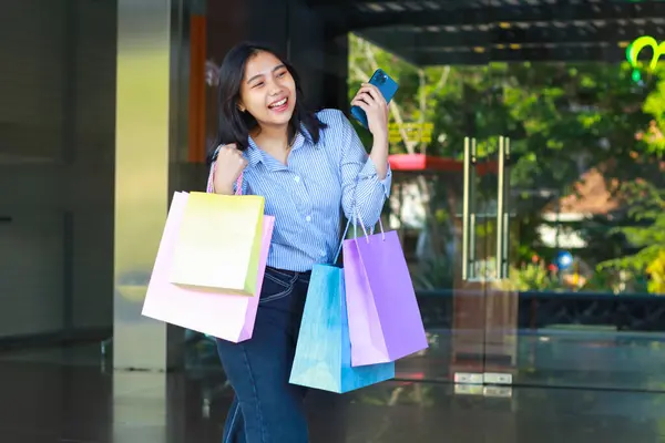 laughing asian woman holding shopping bag and smartphone walking in outdoors around city mall