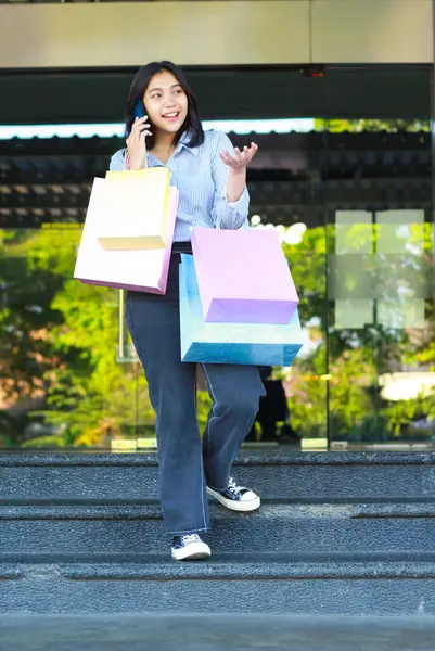 vertical shoot of smiling asian woman holding shopping bag talking on mobile phone while walking on staircase in front of city mall entrance in outdoors