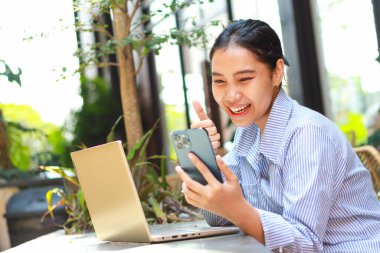 Heyecanlı Asyalı kadın elinde akıllı telefonuyla dışarıda bir kafede dizüstü bilgisayarla otururken elini kaldırıyor.