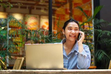 Asyalı heyecanlı bir kadın cep telefonuyla konuşuyor. Laptopuyla dışarıda yeşil bitkilerle dolu bir kafede çalışıyor.