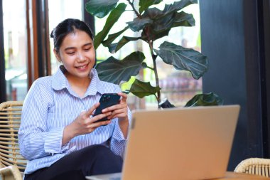 Asyalı heyecanlı kadın yeşil bitkilerle dolu açık bir kafeden çalışırken bilgisayarını ahşap masaya koymuş akıllı telefon kullanıyor.