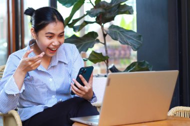 heyecanlı Asyalı kadın ahşap masada dizüstü bilgisayarla konuşuyor ve yeşil bitkilerle birlikte açık kafede çalışırken elini kaldırıyor.