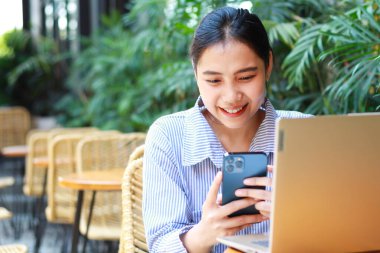 heyecan verici Asyalı kadın laptopuyla ahşap masada yeşil bitkilerle birlikte açık kafeden çalışıyor.