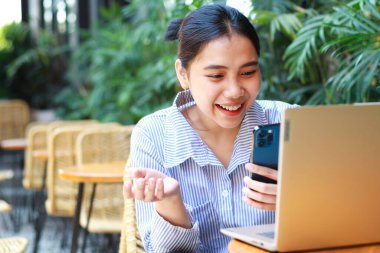 heyecan verici Asyalı kadın laptopuyla ahşap masada yeşil bitkilerle birlikte açık kafeden çalışıyor.