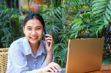 heyecanlı Asyalı kadın dışarıda kafede oturmuş akıllı telefondan konuşuyor ve bilgisayarla çalışırken kameraya bakıp gülümsüyor.