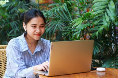 Mutlu Asyalı bir kadın açık hava kafede oturmuş dizüstü bilgisayarla uzaktan kumandalı yeşil gömlekli ve arka planda sıra sıra sandalyelerle çalışıyor.