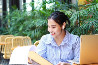 Mutlu Asyalı kadın açık havada oturmuş elinde dizüstü bilgisayarla ahşap masada çizgili mavi gömlekli evrak işleriyle