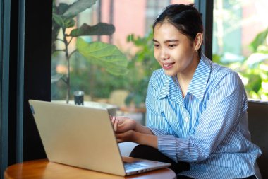 Mutlu Asyalı kadın laptopta çalışırken yeşil bitkilerin olduğu bir kafede oturmaktan zevk alıyor.