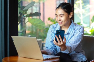 Akıllı telefon tutan mutlu Asyalı kadın ve dizüstü bilgisayar üzerinde çalışırken kafeden online toplantı yapıyorlar.