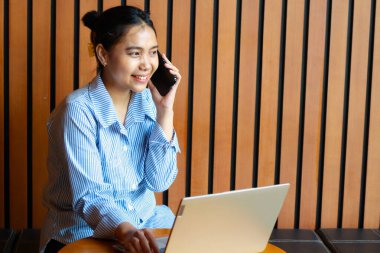 Mutlu Asyalı kadın cep telefonuyla konuşuyor ve laptopla çalışıyor. Kafe kanepede, ahşap arka planda oturuyor.