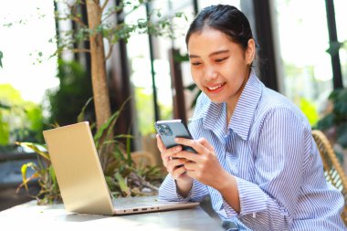 Mutlu Asyalı kadın sosyal medyada geziniyor ve haber okuyor. Masasında dizüstü bilgisayarı olan akıllı telefon kullanarak yeşil bitkilerin olduğu bir kafeden uzaktan çalışıyor.