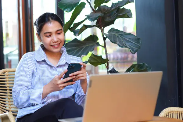 Asyalı heyecanlı kadın yeşil bitkilerle dolu açık bir kafeden çalışırken bilgisayarını ahşap masaya koymuş akıllı telefon kullanıyor.