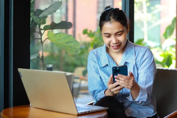 Mutlu Asyalı kadın elinde akıllı telefonuyla sosyal medyada geziniyor ve ahşap masada dizüstü bilgisayarla haber okuyor. Kafeden internet üzerinden toplantı yapıyorlar.