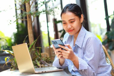 Mutlu Asyalı kadın sosyal medyada geziniyor ve haber okuyor. Masasında dizüstü bilgisayarı olan akıllı telefon kullanarak yeşil bitkilerin olduğu bir kafeden uzaktan çalışıyor.