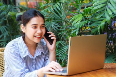 Akıllı telefondan konuşan ve açık kafede laptopla çalışan gülen Asyalı kadın.