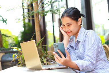 Gülümseyen Asyalı kadın akıllı telefona bakıyor. Mutlulukla düşünüyor. Açık kafede dizüstü bilgisayarla çalışıyor.