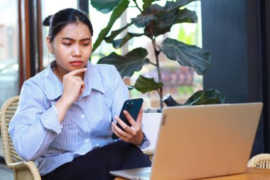 Akıllı telefonlara bakan düşünceli Asyalı kadın çenesinde ellerle dışarıda bir kafede dizüstü bilgisayarla çalışırken ciddi bir şekilde düşün.