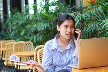 Asyalı iş kadını, açık kafede bilgisayarla çalışırken akıllı telefondan konuşurken evrak işi raporunu tutarken kafası karışmış görünüyor.