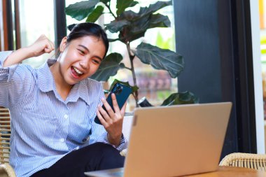 Akıllı telefona bakan ve yumruğunu kaldıran başarılı Asyalı kadın, zaferi ve başarıyı kafede dizüstü bilgisayarla çalışırken kutluyor.