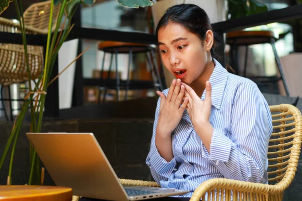pretty asian business woman shocked looking at laptop while working remotely in outdoor cafe covering her open mouth with hands show wow expression