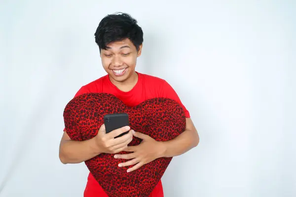 laughing asian man hug heart shape pillow while holding smartphone isolated on white background
