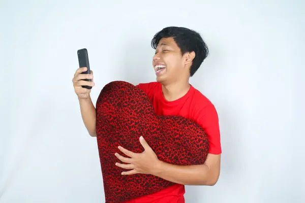 laughing asian man hug heart shape pillow while holding smartphone isolated on white background