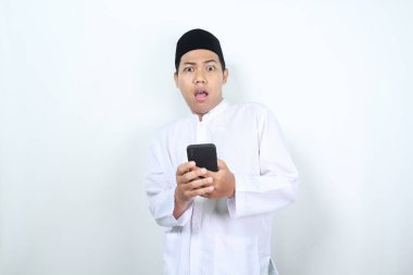 Şok olmuş Asyalı Müslüman adam elinde cep telefonuyla kameraya bakıyor.