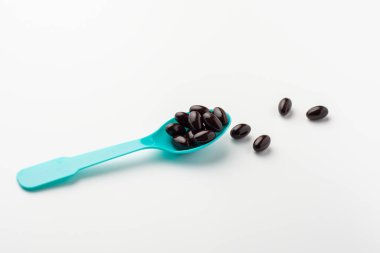 Beyaz arka planda mavi kaşıkta siyah yumuşak kapsüller. Vitamin takviyesi