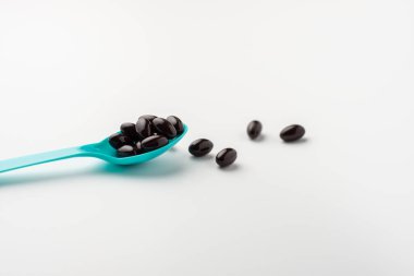 Beyaz arka planda mavi kaşıkta siyah yumuşak kapsüller. Vitamin takviyesi