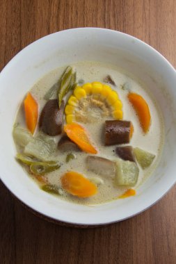 Sayur lodeh ya da sebze soslu hindistan cevizi sütü, kahverengi ahşap bir masada servis edilen beyaz kasede bulunan geleneksel bir Endonezya mutfağı yemeğidir..