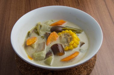 Sayur lodeh ya da sebze soslu hindistan cevizi sütü, kahverengi ahşap bir masada servis edilen beyaz kasede bulunan geleneksel bir Endonezya mutfağı yemeğidir..