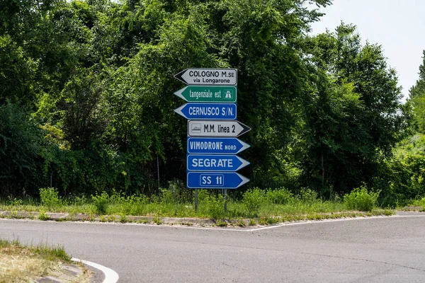 Şehirleri, sokakları ve metroyu gösteren tabelalar. Arka planda ağaçlar olan bir banliyö yolunun kenarında.