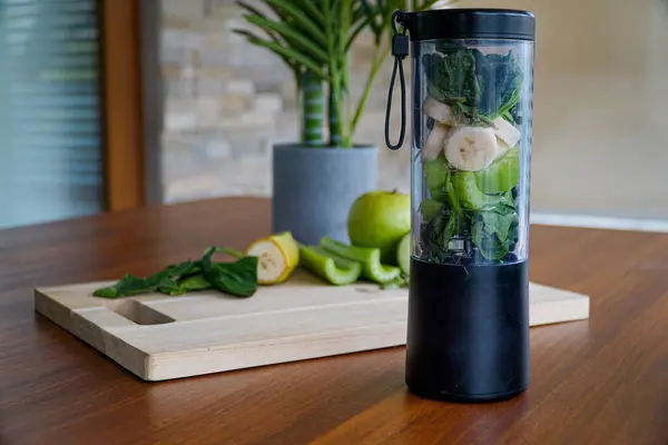 A green detox smoothie in the making! A juicer is loaded with organic spinach, ripe banana, crisp celery, and juicy apple. Behind it, a rustic wooden cutting board showcases these fresh ingredients