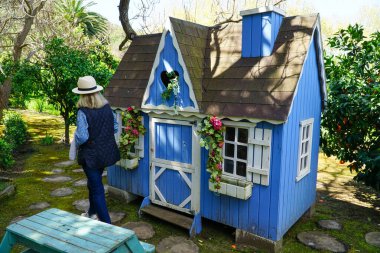 An eager grandmother waits to play with her grandchildren in the charming garden beside the blue wooden dollhouse clipart