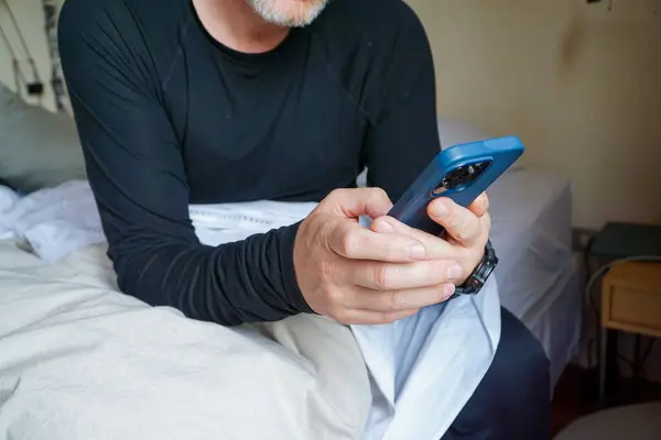 An unrecognizable 60-year-old man is seen looking at his phone from the bed.