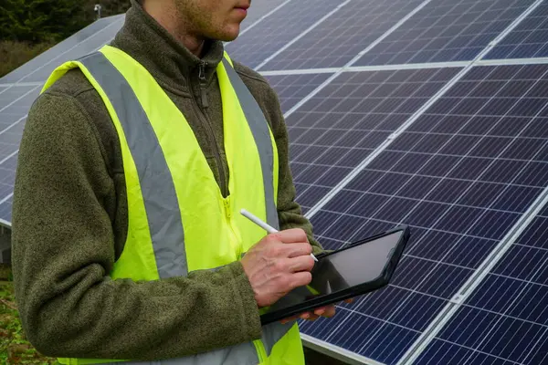 A young environmental engineering student learning about renewable energy while examining solar panels and jotting down notes on his tablet.