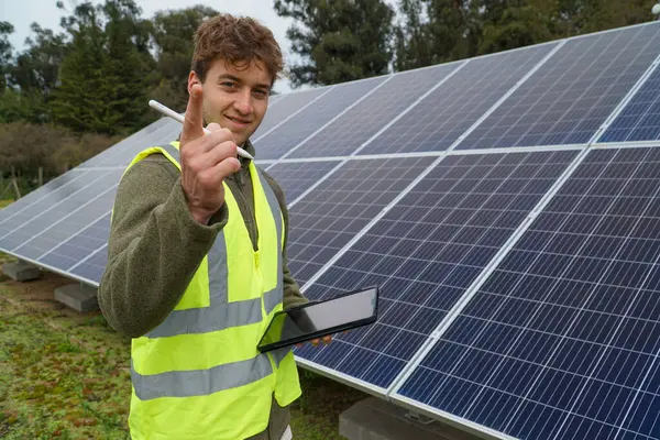 A young engineer smiling at the camera while monitoring solar panels on his tablet on a cloudy day.