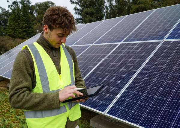 A young engineer writing on his tablet while monitoring solar panels.