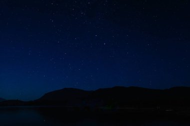 Gece vakti, yıldızlarla süslenmiş koyu mavi bir gökyüzü, dağ çizgisi ve yansıtıcı bir göl..