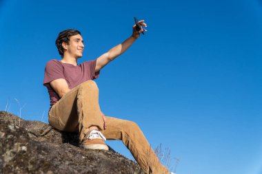 Genç adam bir kayanın üzerinde oturuyor, kameraya gülümseyen bir anı yakalıyor..