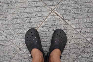 Çapraz oyukları olan desenli beton bir kaldırımda duran siyah takunyalı ayakların yakın çekimi..