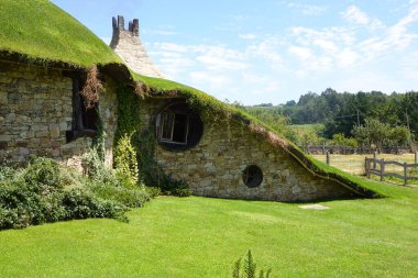 Çimen kaplı çatı ile büyüleyici bir hobbit tarzı kulübe verimli bir kırsal alana yerleşmiş, yuvarlak pencereler ve taş duvarlar içeriyor..