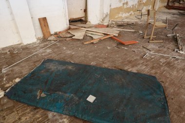 A derelict school gym with a torn blue mat on the floor, surrounded by broken furniture and scattered debris, reflecting neglect and decay. clipart