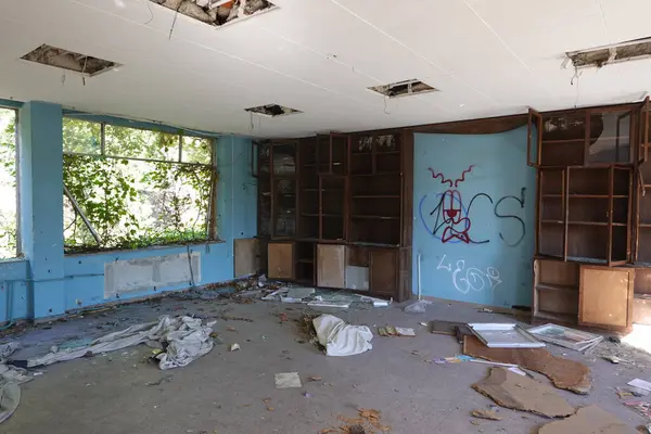 stock image An abandoned classroom with broken ceiling tiles, open shelves, graffiti on the walls, and windows overtaken by vines.