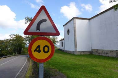 Kavisli trafik levhası ve saatte 40 km hız sınırı işareti beyaz bir binanın yanındaki kırsal bir yolun yanında duruyor. Kırmızı üçgen uyarı işareti açık mavi gökyüzü ve arka planda yeşillik ile keskin bir sağ dönüş olduğunu gösteriyor..