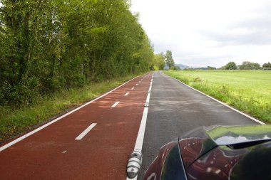 Long road with separate bike lane bordered by dense green trees on one side and open fields on the other under a cloudy sky. A red bike path runs parallel to the road, separated by white lines and small dividers. clipart