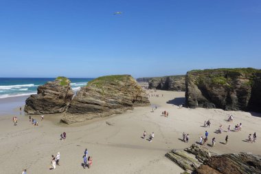 Playa de las Catedrales, İspanya 'da güneşli bir günde kumlu bir plaj boyunca büyük kayalık oluşumlarla yürüyen turistler. Açık mavi gökyüzü ve kayalara çarpan dalgalar kıyı manzarasını güzelleştirirken, bir grup insan benzersiz bölgeyi keşfediyor.