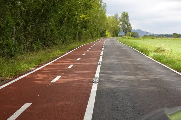 Bitişiğinde kırmızı bisiklet yolu olan boş kırsal yol, küçük yansıtıcı ayırıcılarla ayrılmış. Yol, bir taraftaki yeşil ağaçların arasından diğer taraftaki açık arazilere doğru uzanıyor, parçalı bulutlu bir gökyüzünün altında..