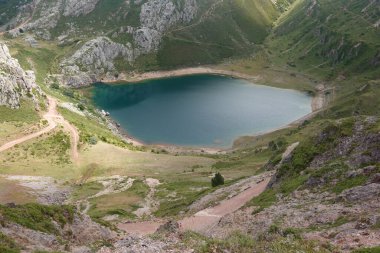 Somiedo Doğal Parkı, Asturias 'taki La Cueva Gölü' nün yukarıdan görünüşü. Bereketli yeşil tepeler ve engebeli manzaralarla çevrili sakin mavi sulara uzanan dolambaçlı toprak bir patika ve kayalık uçurumlar..